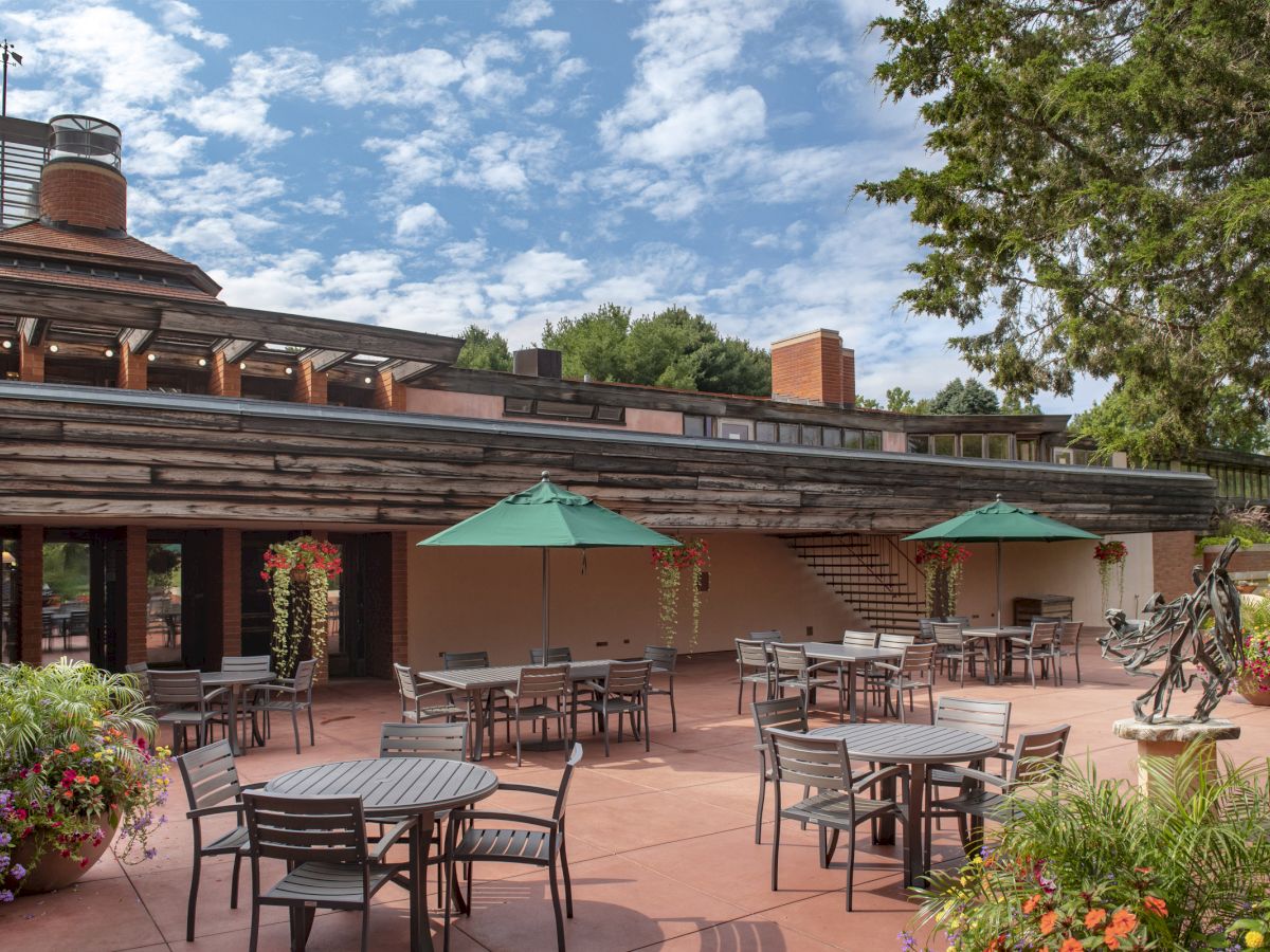 The image shows an outdoor patio with tables, chairs, and umbrellas in front of a modern building surrounded by greenery and flowers.