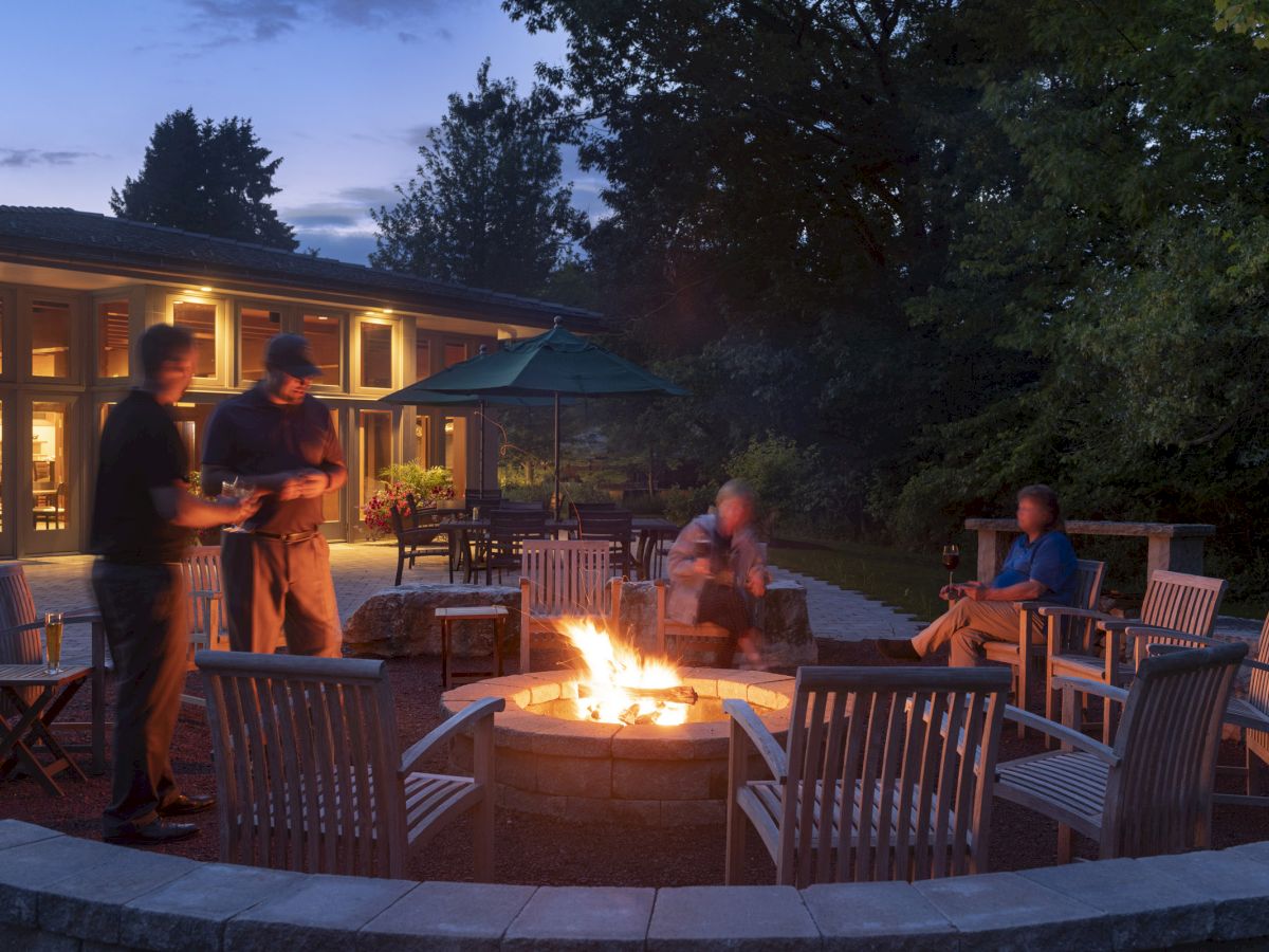 People gathered around a fire pit in a patio area, with outdoor seating and trees in the background. Evening atmosphere captured.