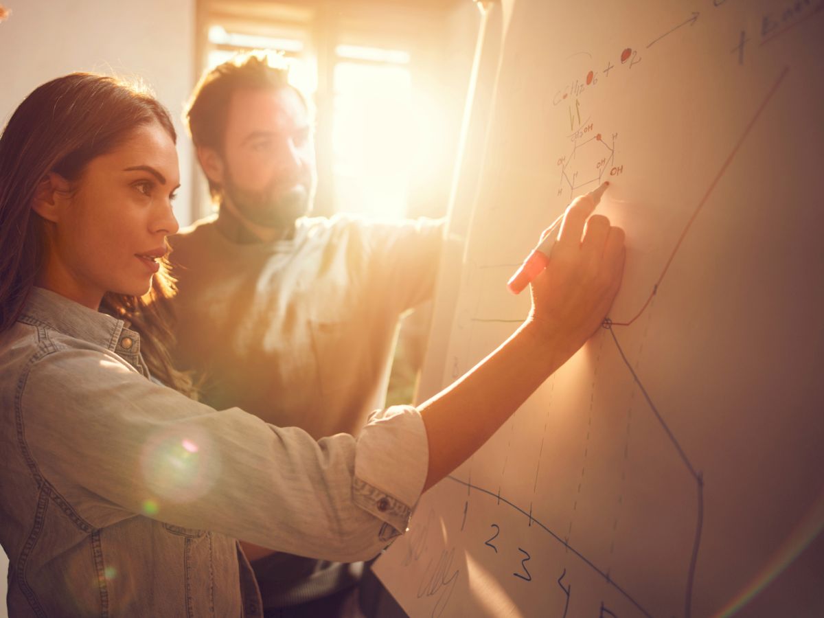 Two people are discussing data on a whiteboard, with sunlight streaming through a window in the background.