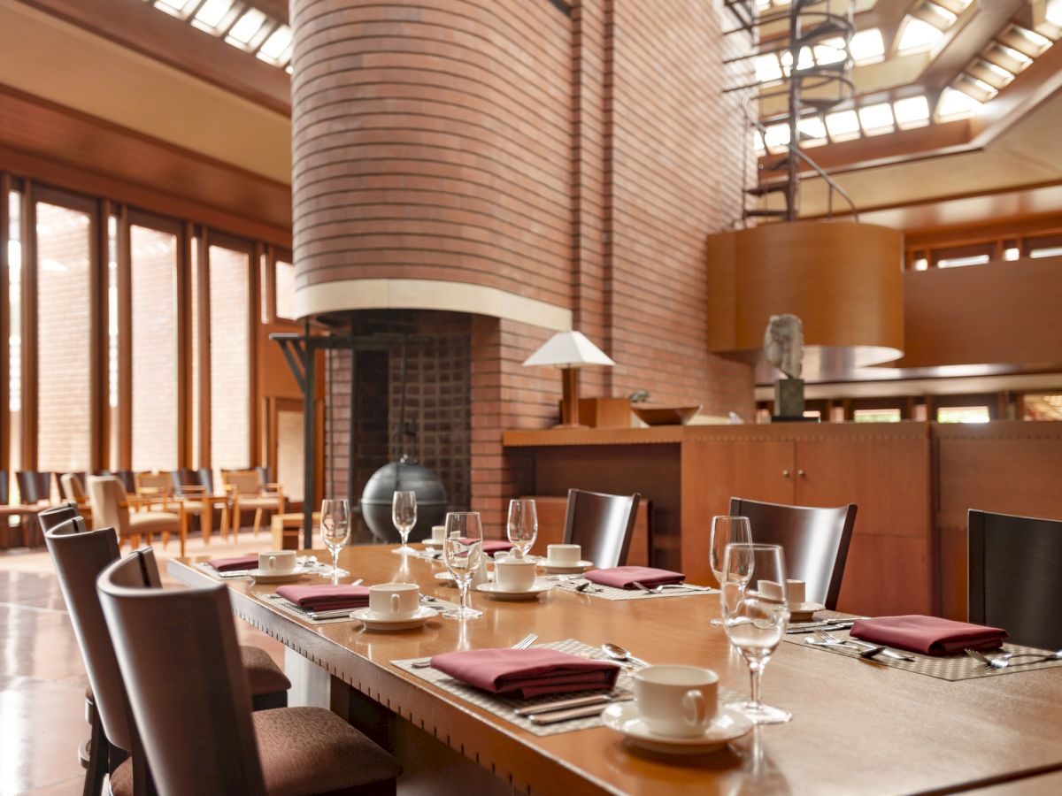 A modern dining area with a long table, set with glassware and napkins, features a unique brick pillar and abundant natural light.