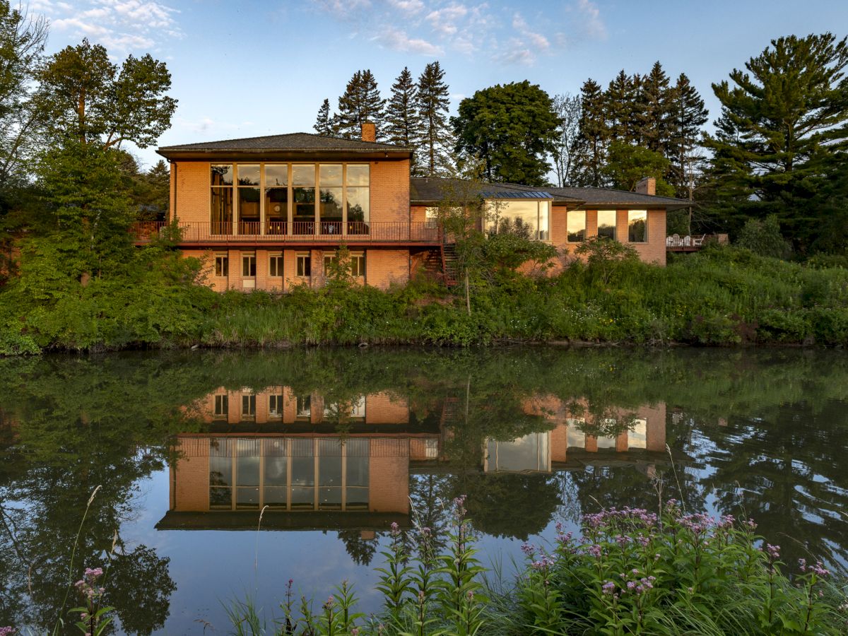 A modern house with large windows sits by a calm river, surrounded by lush greenery and trees, reflecting beautifully in the water.
