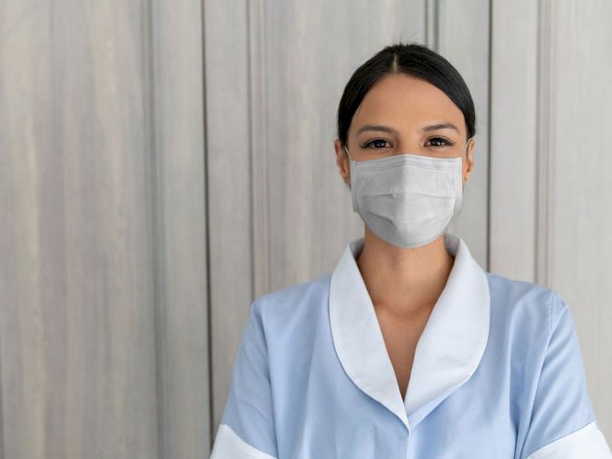 A person wearing a face mask and a light blue uniform stands in front of a wooden background, facing the camera directly.