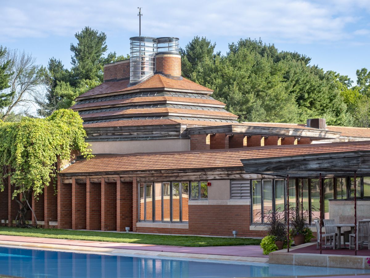 The image shows a building with unique architecture, featuring a tiered roof and large windows, set amidst greenery and a pool.