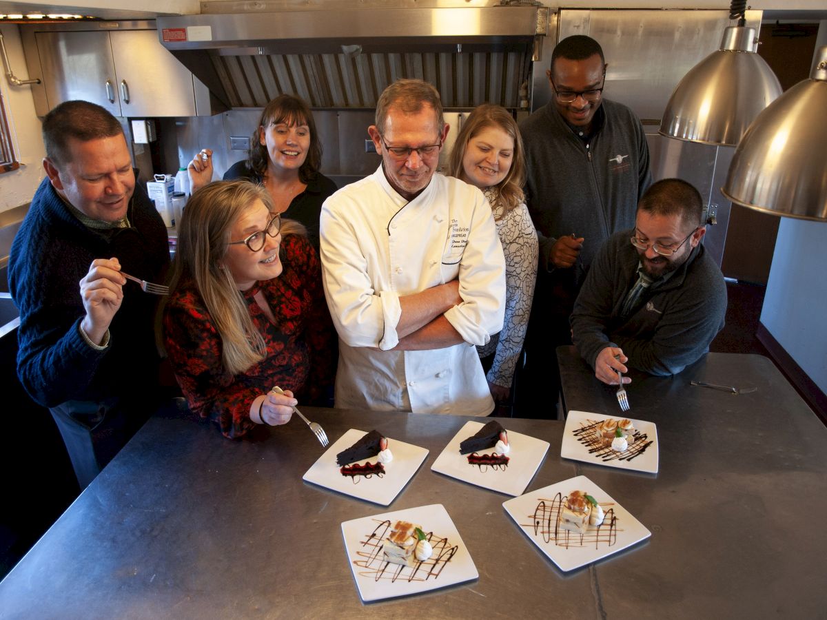 A chef stands with a group of people in a kitchen, presenting four plated desserts. The group looks eager to taste the dishes.