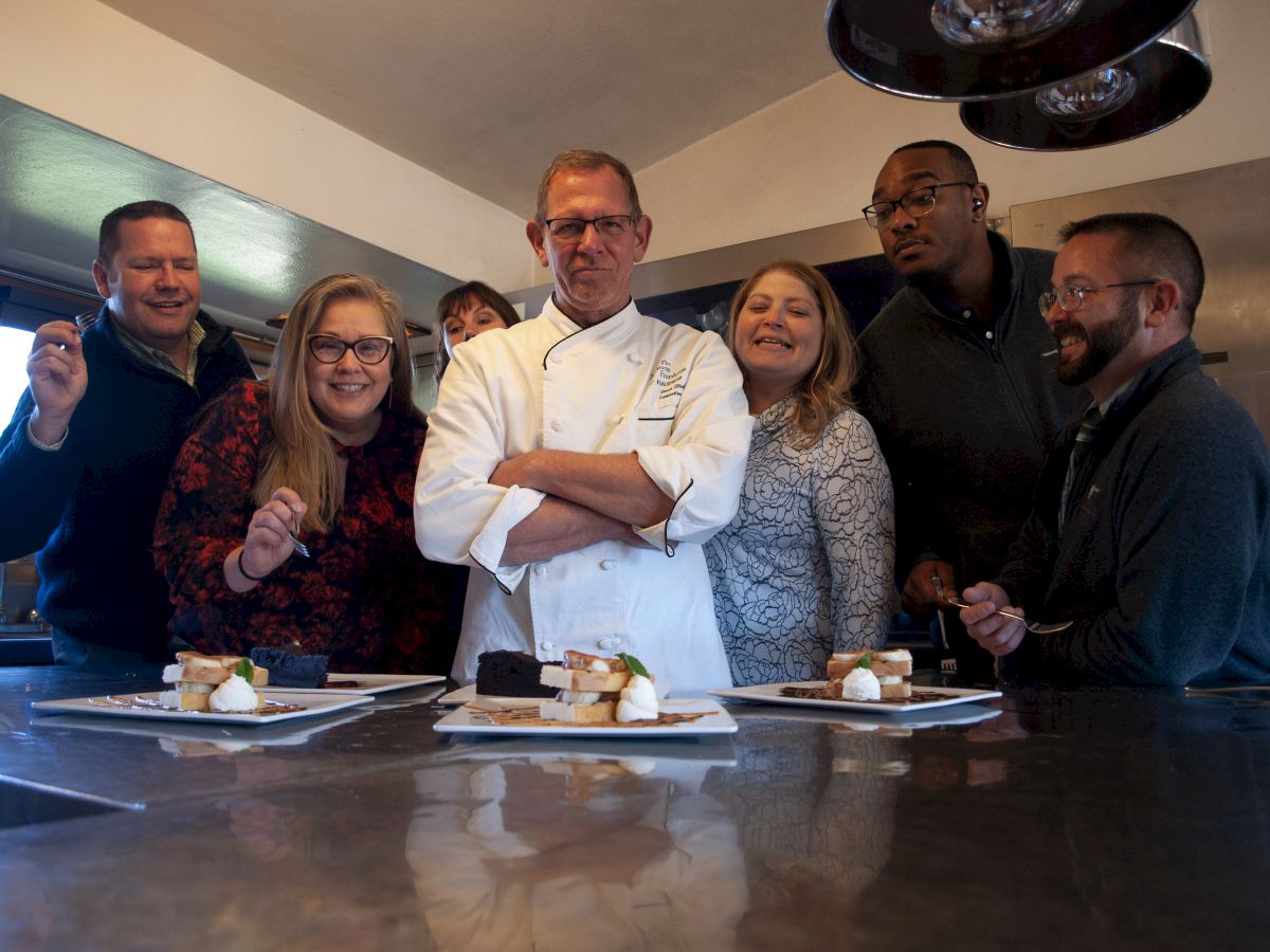 A group of people is gathered around a chef, who is standing confidently with arms crossed. Plates of dessert are in front of them on a table.