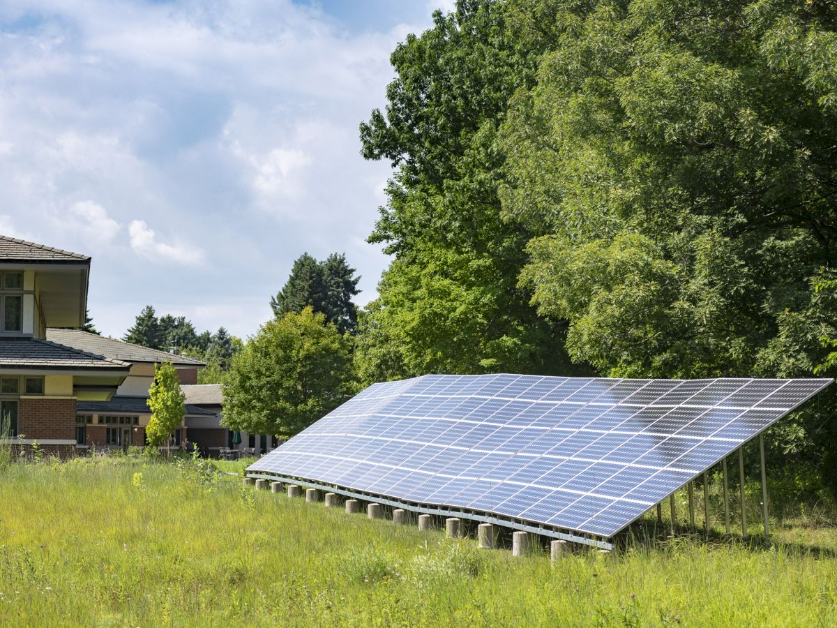 A solar panel array is positioned in a grassy area beside a building, surrounded by trees and under a partly cloudy sky.
