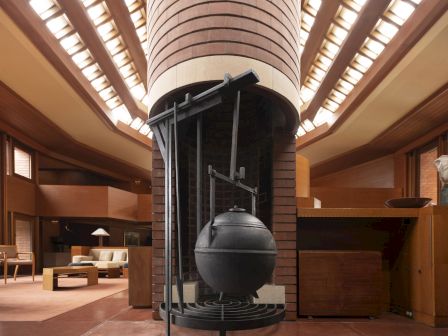 The image shows a large, unique fireplace with a hanging metal kettle in a spacious room with high ceilings and rows of windows.