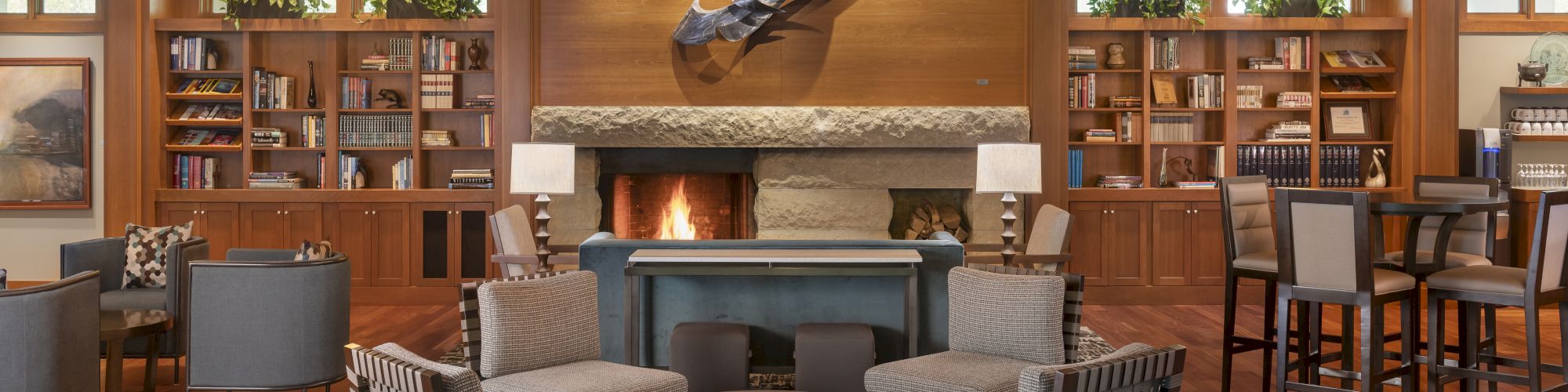 A cozy lounge with a fireplace, armchairs, a rug, bookshelves, and a sculptural wall decoration, creating a warm and inviting atmosphere.