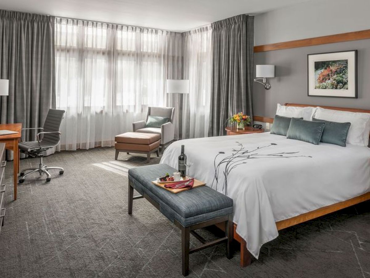 A modern hotel room with a king-sized bed, desk, armchair, and decor. It looks bright with large windows and stylish furnishings.