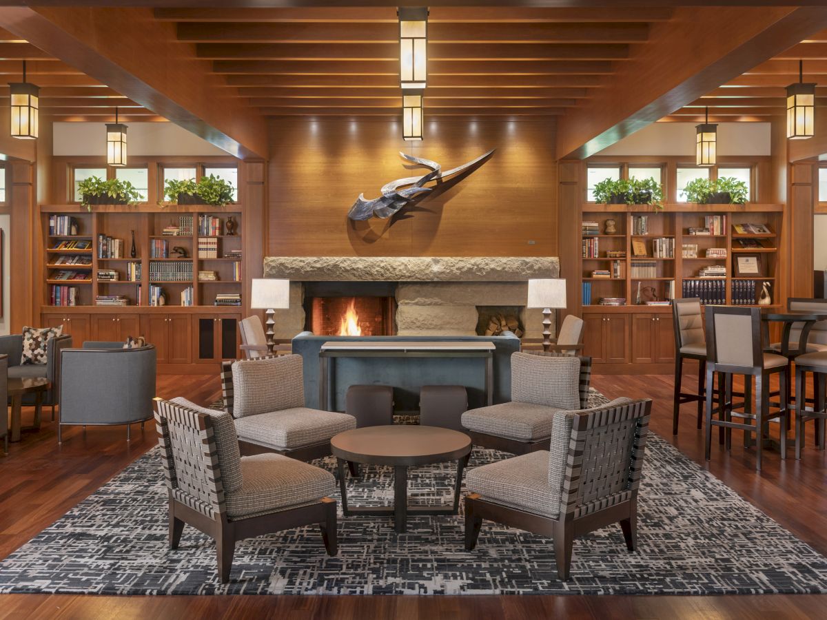 A cozy lounge with armchairs, a fireplace, bookshelves, and plants, featuring warm lighting and wooden accents, creating a welcoming atmosphere.