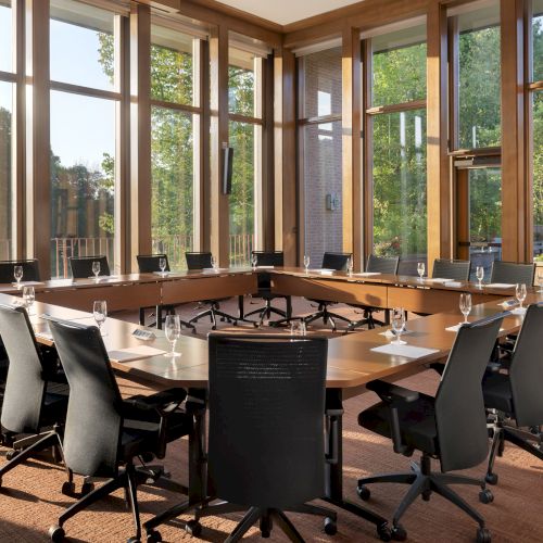 A spacious conference room with a U-shaped table, several chairs, and large windows showing greenery, creating a bright atmosphere.