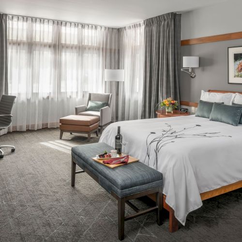 A modern hotel room with a large bed, gray curtains, a bench at the foot, a seating chair by the window, a desk and chair, lamps, and wall art.