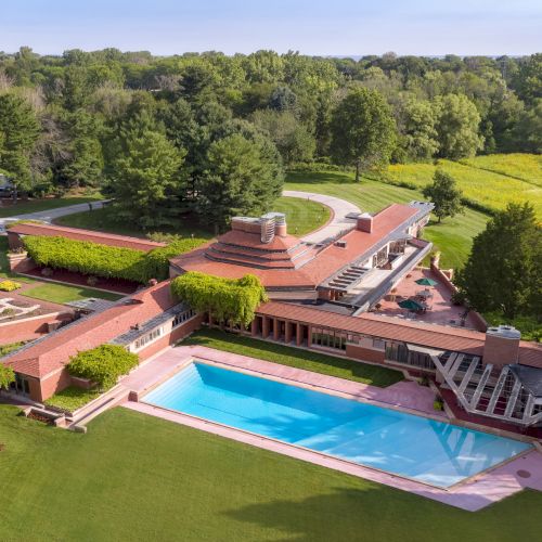 An aerial view of a large modern house with a unique design, adjacent swimming pool, and surrounded by lush greenery and trees.