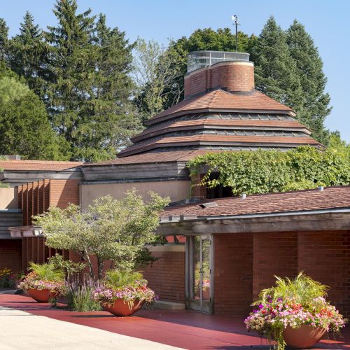 A building with a unique, tiered roof surrounded by greenery and potted plants, featuring red brick walls and a clear blue sky.