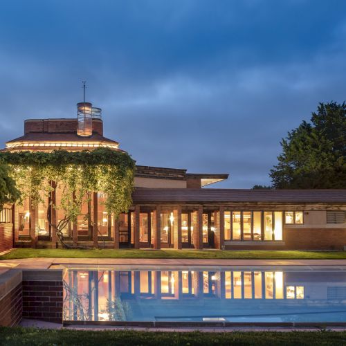 The image shows a modern building with large windows, surrounded by trees, next to a pool reflecting the structure.