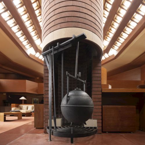 The image shows a spacious room with a central, modern fireplace featuring a large hanging cauldron beneath a tall, cylindrical chimney.