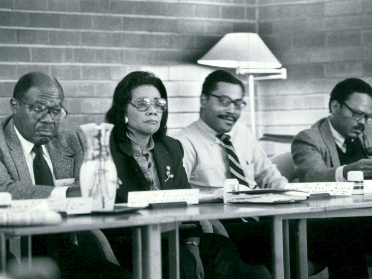 A black and white photo shows four people sitting at a table in a meeting setup, with papers and a lamp in the background.