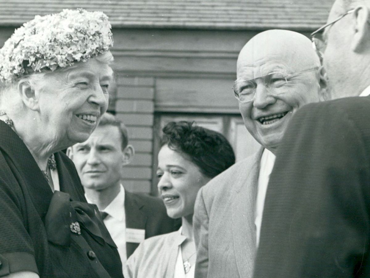 A group of people in formal attire are conversing happily outdoors, with one woman wearing a floral hat.