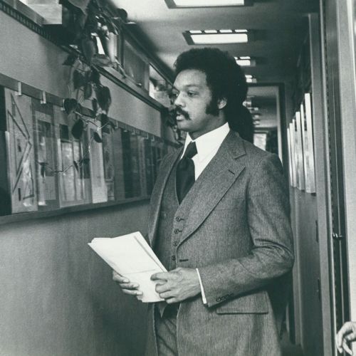 A man in a suit holds papers while walking down a hallway lined with framed pictures and plants.