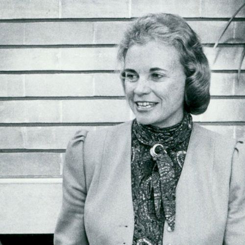 The image shows a woman smiling and talking to another person, standing in front of a brick wall with some greenery.