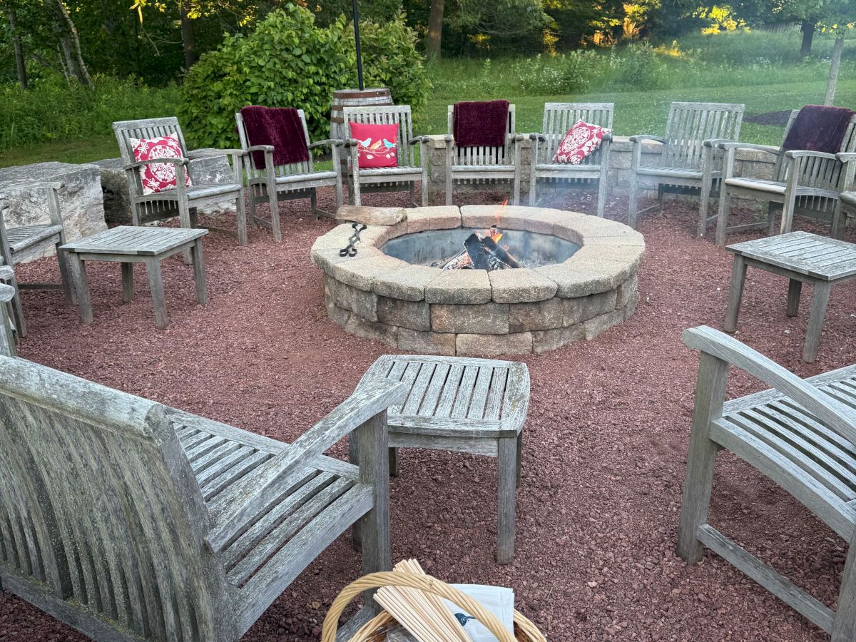 Wooden chairs encircle a stone fire pit with a basket of logs. Greenery surrounds the setup, creating a cozy outdoor gathering spot.