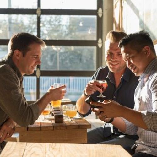 Three people sitting around a table with drinks, smiling, and looking at a phone in a sunlit room.