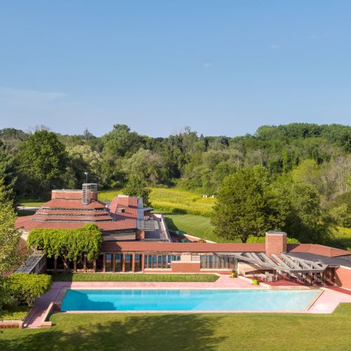 A spacious, modern house with a flat roof is surrounded by greenery. A large rectangular pool is in the foreground.