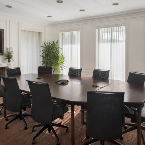 A conference room with a large oval table, several chairs, a portrait on the wall, plants, and natural light streaming through the windows.