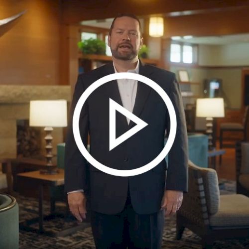 A man in a suit stands in a warmly lit, elegantly furnished living room with modern decor and a large play button overlay in the center.