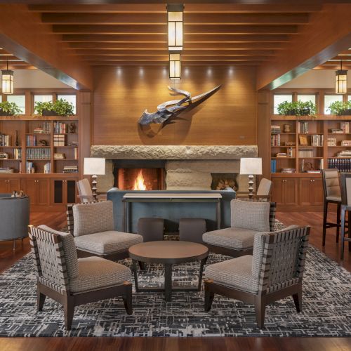 A cozy lounge with chairs around a fireplace, bookshelves, and tables, featuring warm lighting and wooden decor throughout the room.