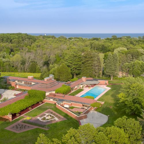 An aerial view of a large, contemporary house with a pool, surrounded by trees, near a body of water.