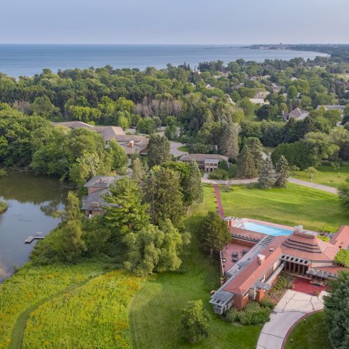 Aerial view of a large estate with gardens, a pond, and architectural buildings amidst lush greenery, near a large body of water.