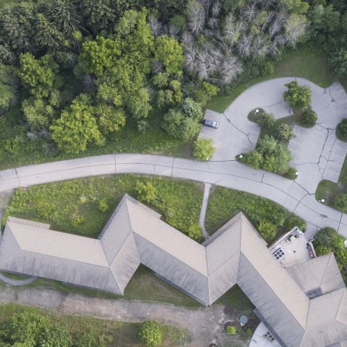 An aerial view of a building surrounded by a road, trees, and greenery, with a driveway leading to the entrance.
