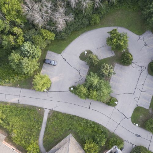 An aerial view of a parking lot with one car, surrounded by trees and intersected by a road with visible cracks in the pavement.