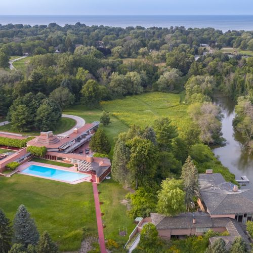 An aerial view of a large, modern house with a pool, surrounded by trees, near a body of water and a lush green landscape.