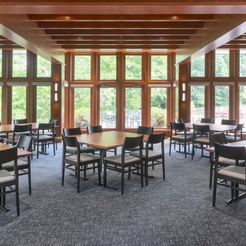 The image shows a dining area with multiple tables and chairs, large windows, and a modern wooden interior design, overlooking greenery.