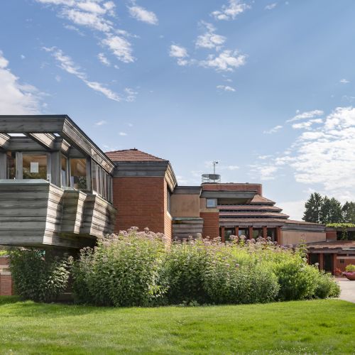A uniquely designed building with brick and metal elements, surrounded by greenery and a partly cloudy sky.