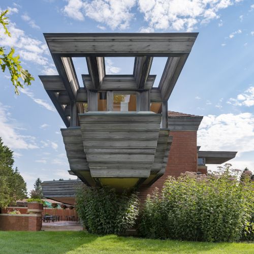 A modern architectural structure with an overhanging design, featuring metal and wood elements, surrounded by greenery and a bright sky.