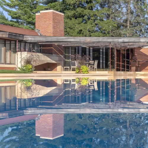 The image shows a modern house with brick and glass architecture, featuring a reflection in a clear blue pool, surrounded by trees.