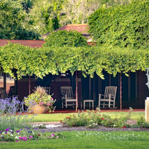 A garden with lush greenery, a vine-covered pergola, outdoor seating, flowers, and a statue on a pedestal can be seen in the image.
