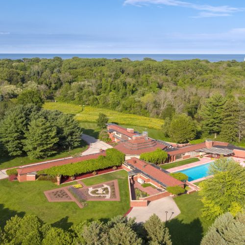 An aerial view of a large, modern home with a swimming pool, surrounded by lush greenery and trees, near a coastline.