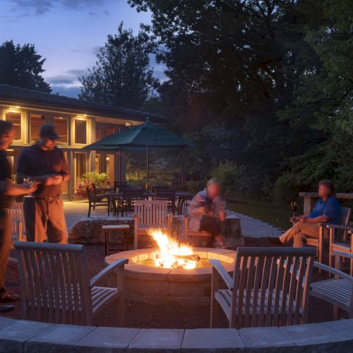 People sitting around a fire pit in a backyard setting with chairs and tables, surrounded by trees and a building at dusk.