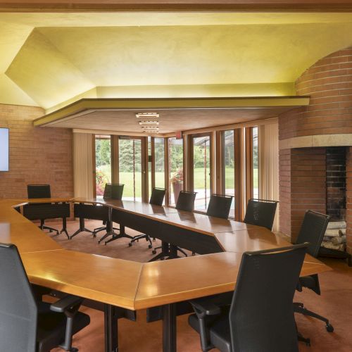 A modern conference room with a unique U-shaped table, chairs, a flat-screen display, brick walls, a fireplace, and large windows.