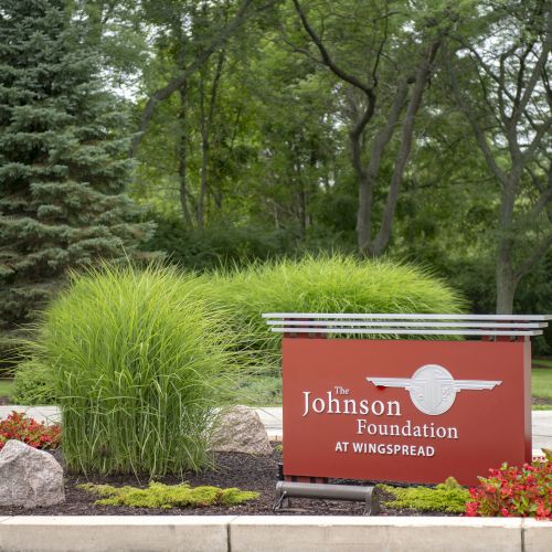 The image shows a sign for "The Johnson Foundation at Wingspread" amidst landscaped greenery and trees in the background.
