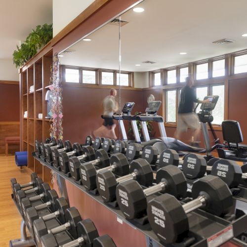 This image shows a gym with dumbbells, treadmills, exercise bikes, and a stability ball, set in a room with wooden floors and mirrors.