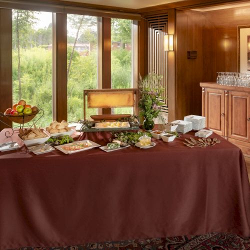 A buffet table with various foods, plates, and a tiered fruit basket is set in a room with wooden furniture and large windows.