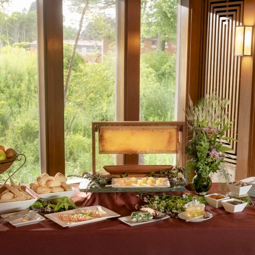 A buffet setup with fruits, bread, salads, and utensils on a table by large windows with a nature view.