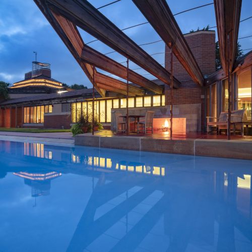 The image shows a modern house with large windows, geometric wooden beams, and a pool reflecting the sky at dusk.