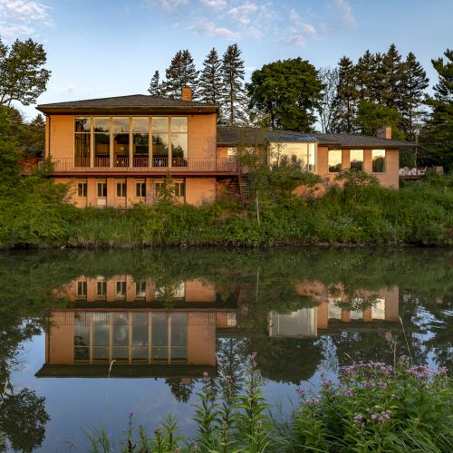 A modern house with large windows is reflected in a calm river, surrounded by lush greenery and trees under a clear blue sky.