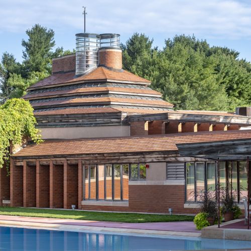 A modern architectural building with layered brick and glass design, surrounded by greenery and a reflective pool in the foreground.